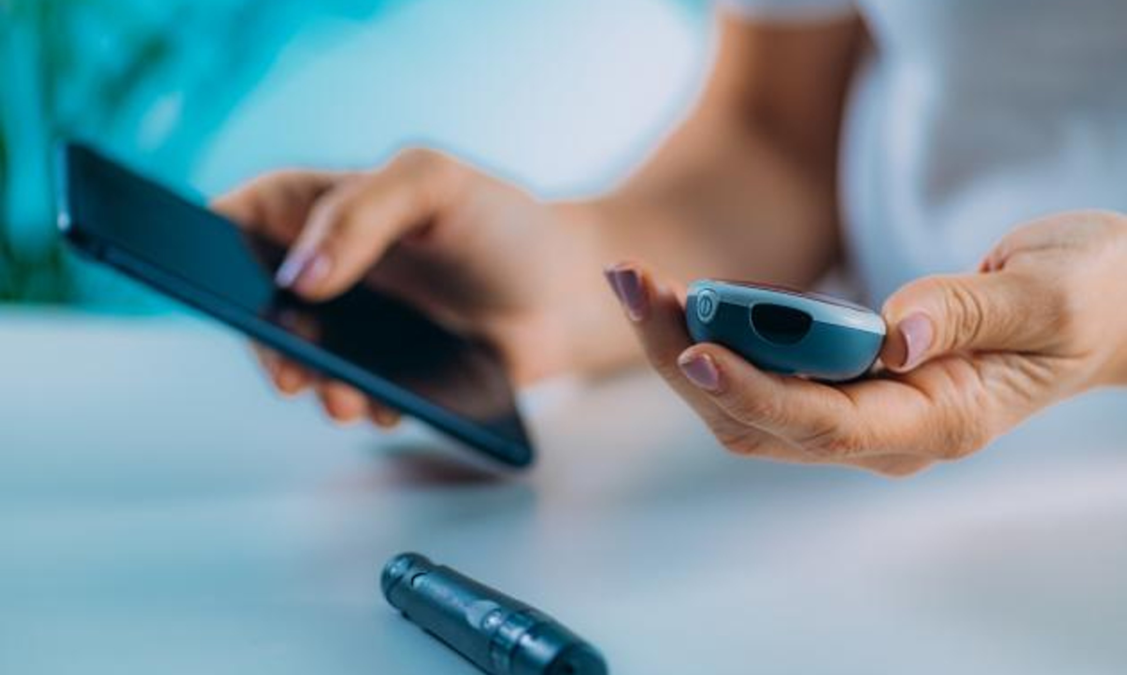 A person holding a glucometer and phone, with a lancet on a desk.
