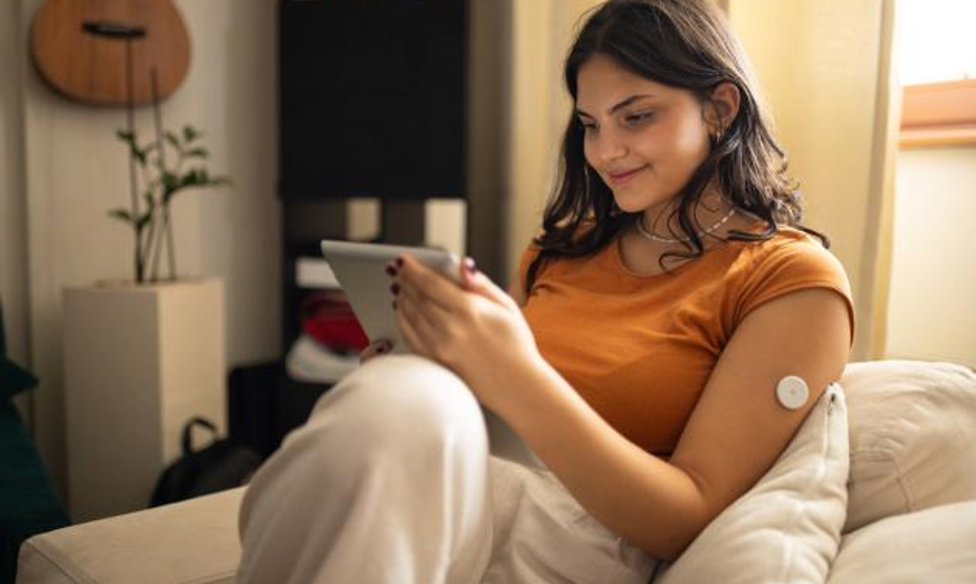 A woman wearing a CGM reviews readings from a tablet while sitting on a couch.