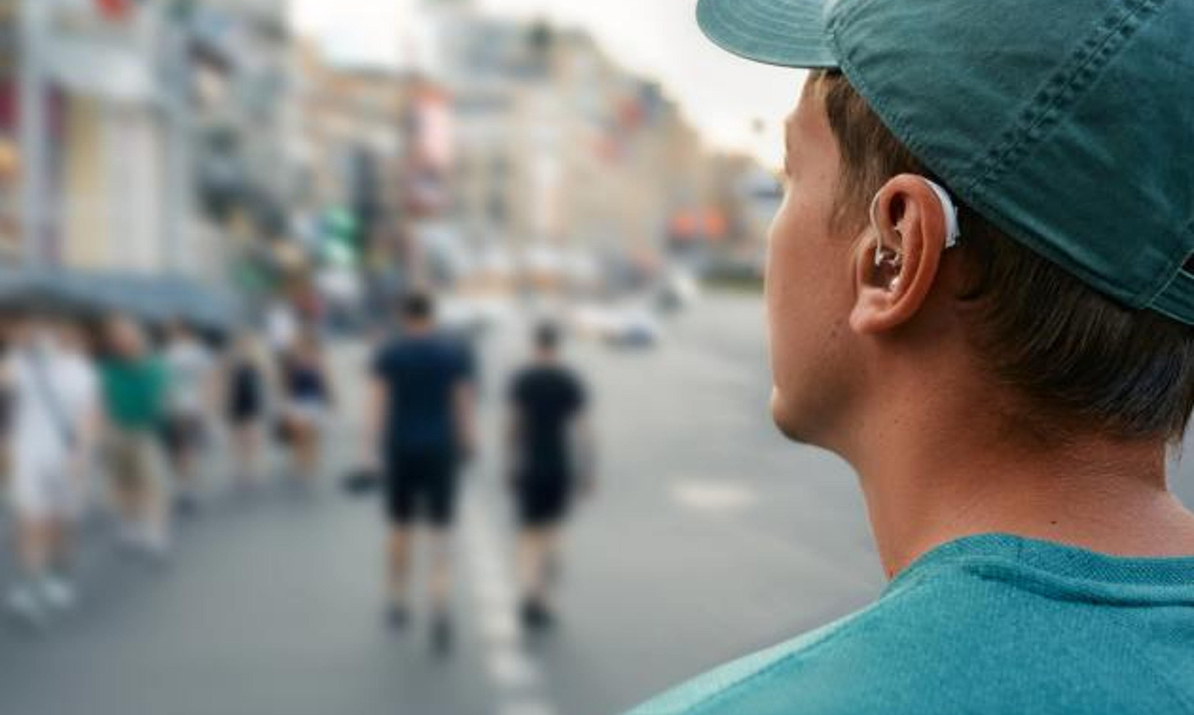 Man with a hearing aid wearing a baseball camp looks on as people stroll down a busy street.