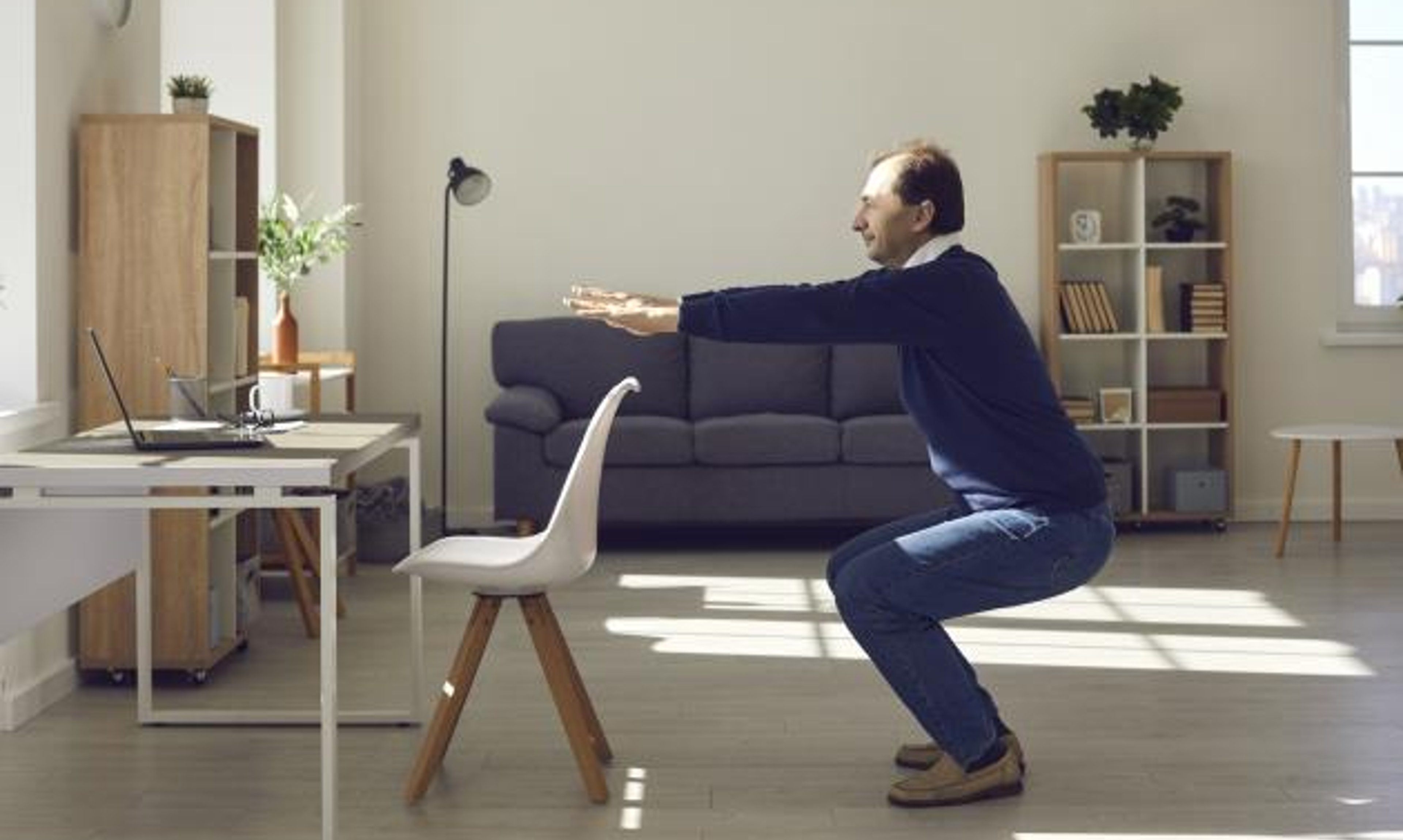 A man at home does a standing squat exercise in front of a chair at a desk.