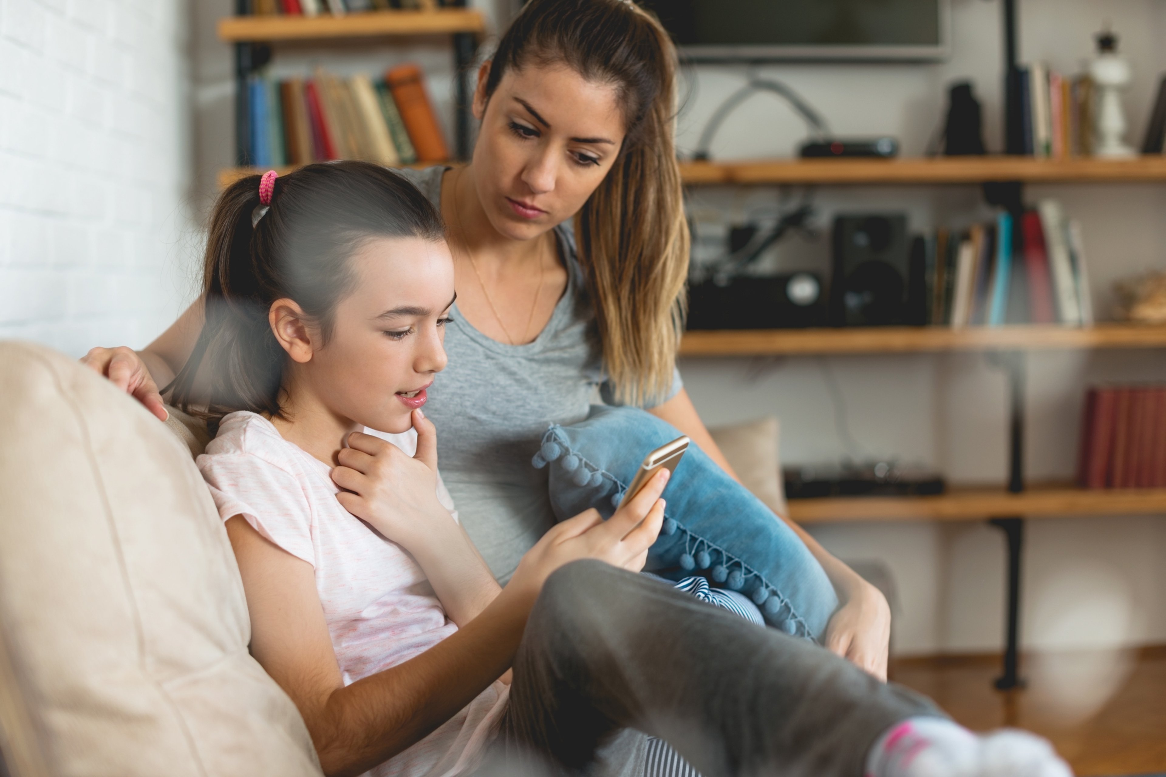 A child holds a smartphone while sitting with her mother on a couch.