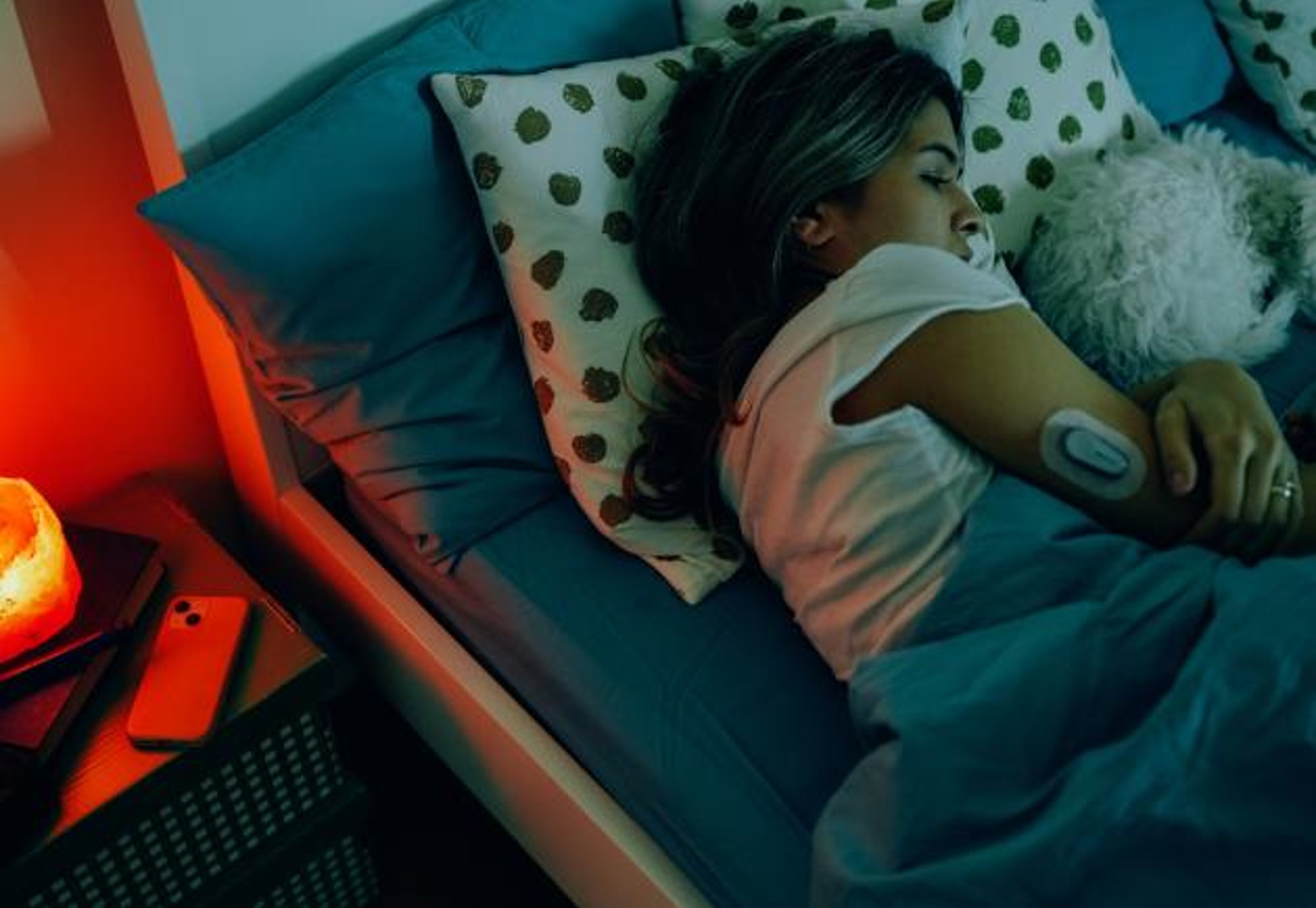 A woman wearging a CGM sensor sleeps next to a soft light on a bedside table.