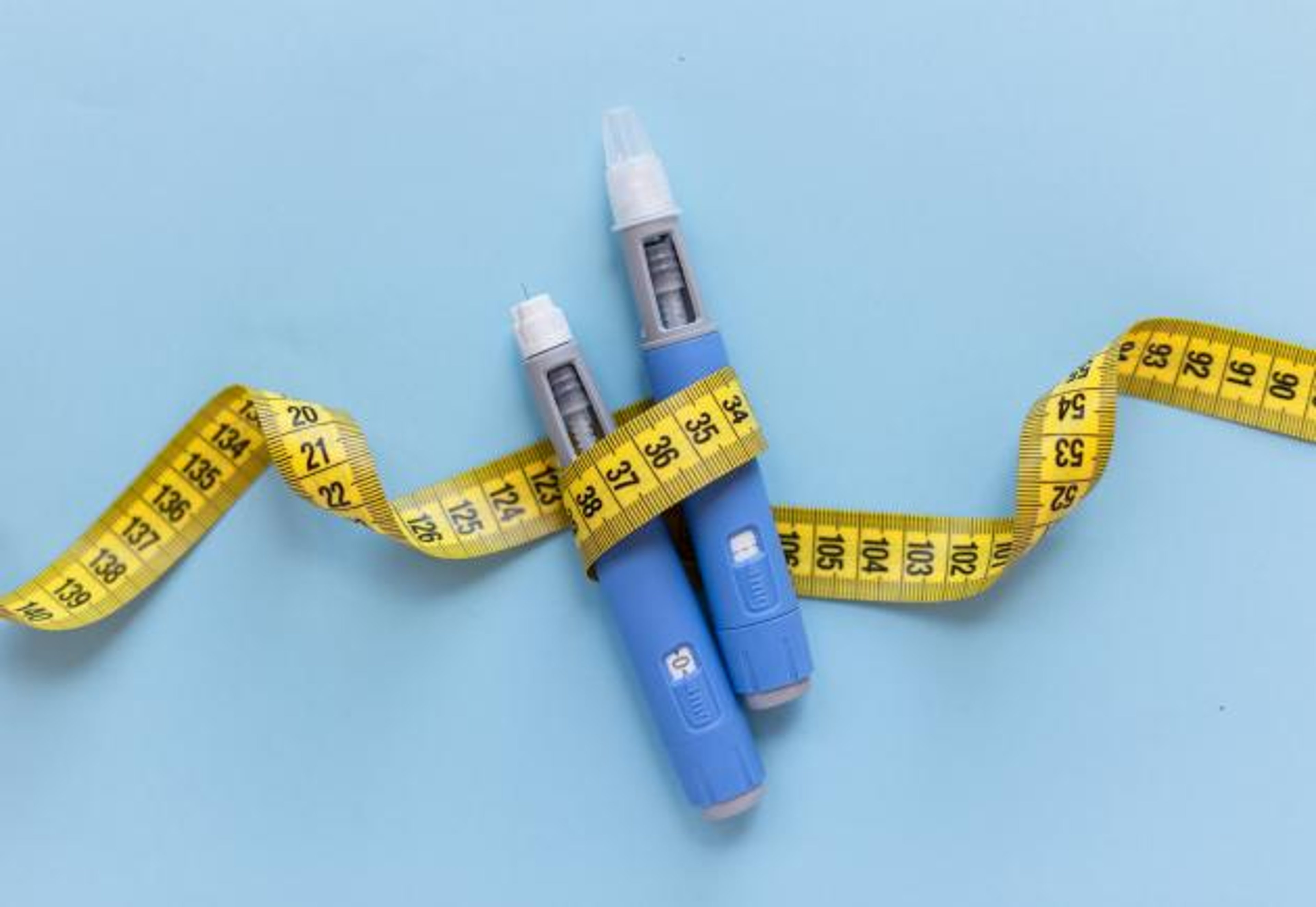Two injectors dosing pens in front of blue background with yellow measuring tape around the injectors.