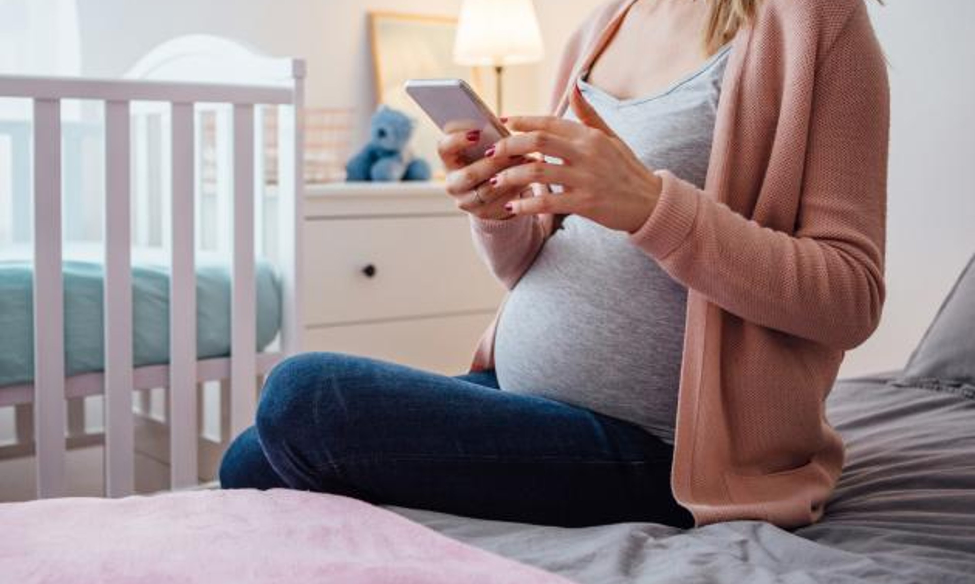 A woman who is pregnant holds a smartphone next to a crib.
