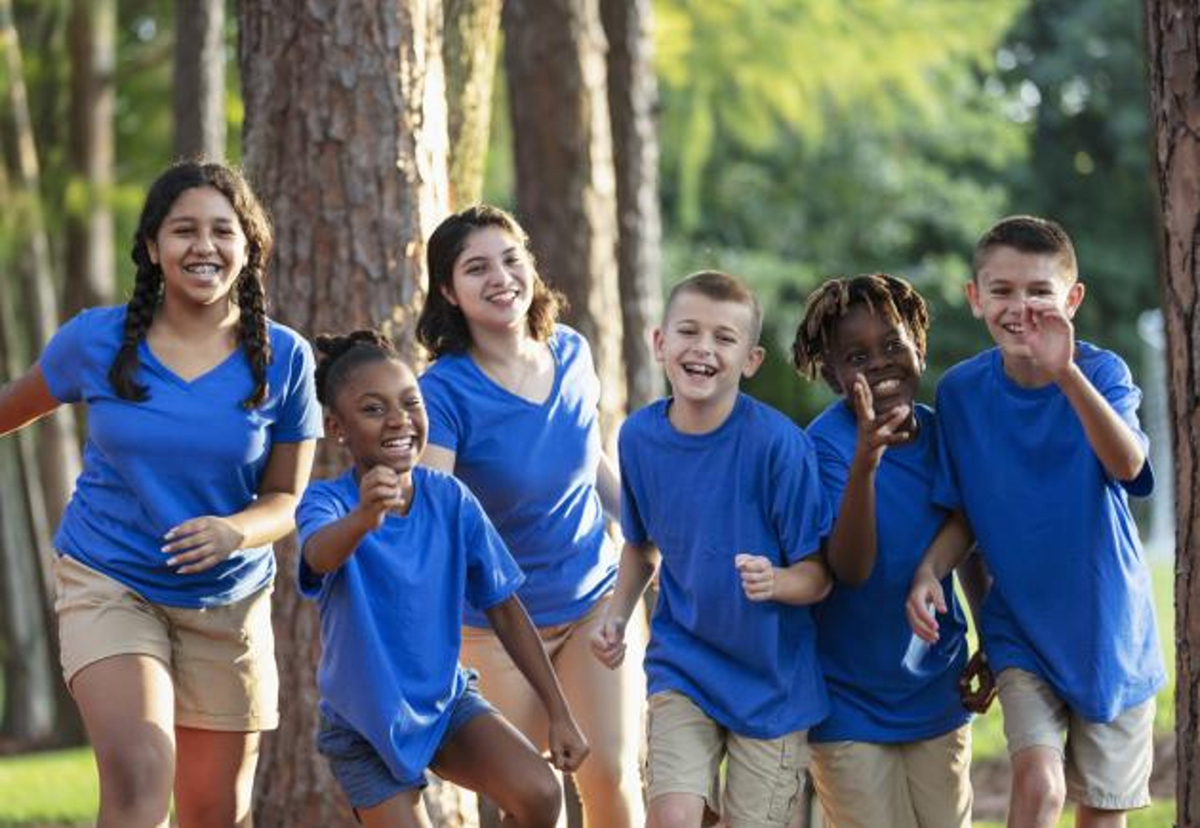 6 children running toward camera wearing camp gear with forest background