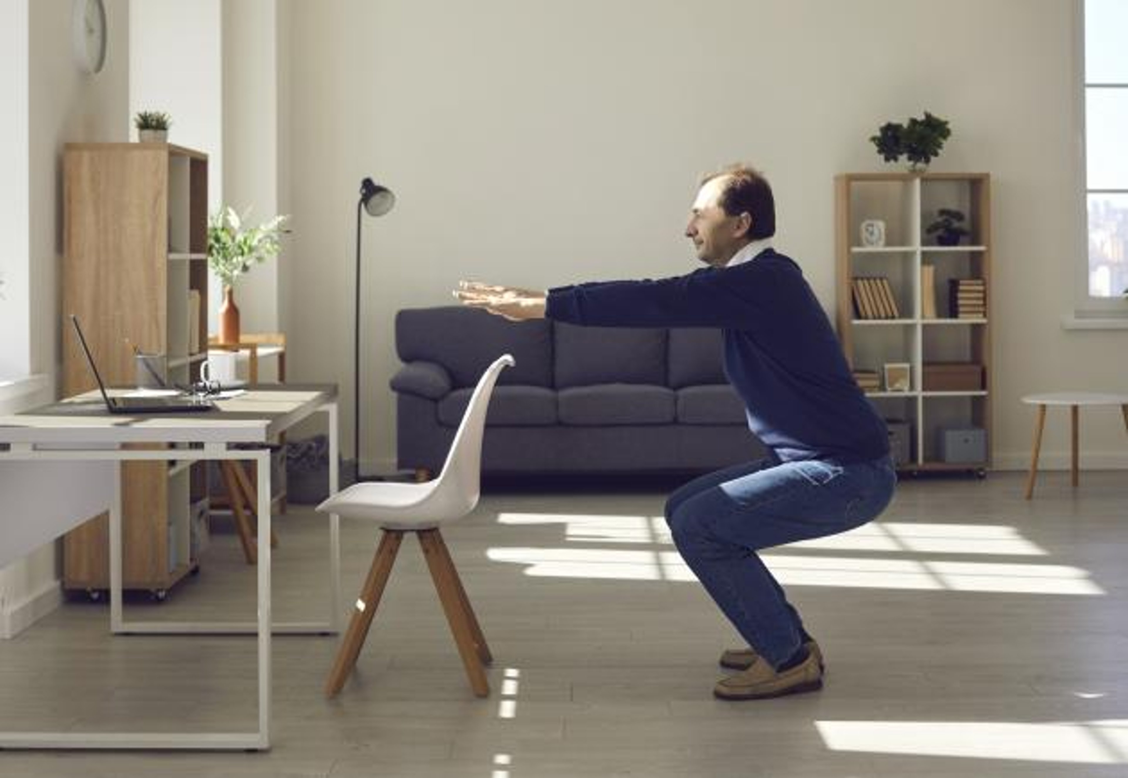 A man at home does a standing squat exercise in front of a chair at a desk.