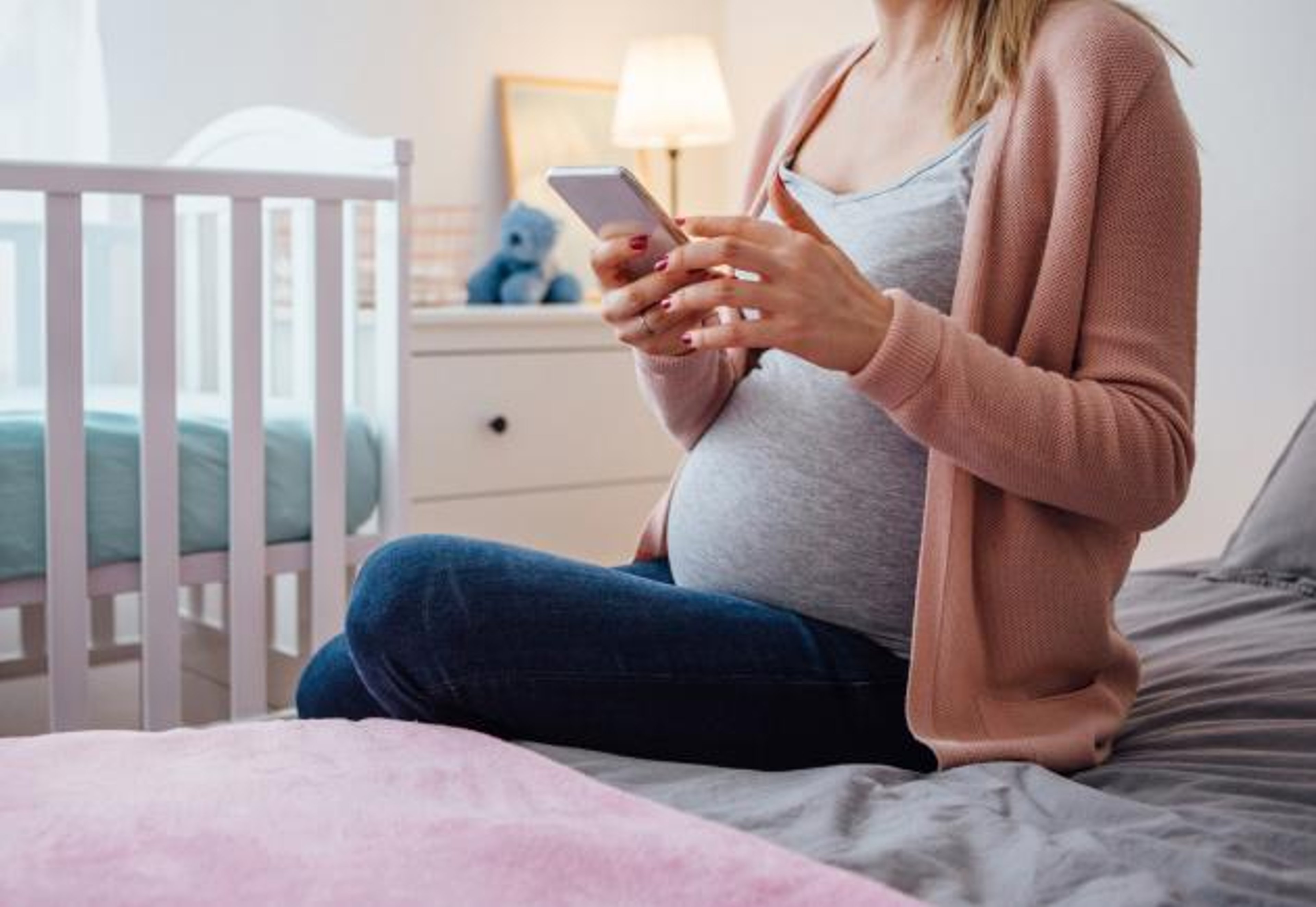 A woman who is pregnant holds a smartphone next to a crib.